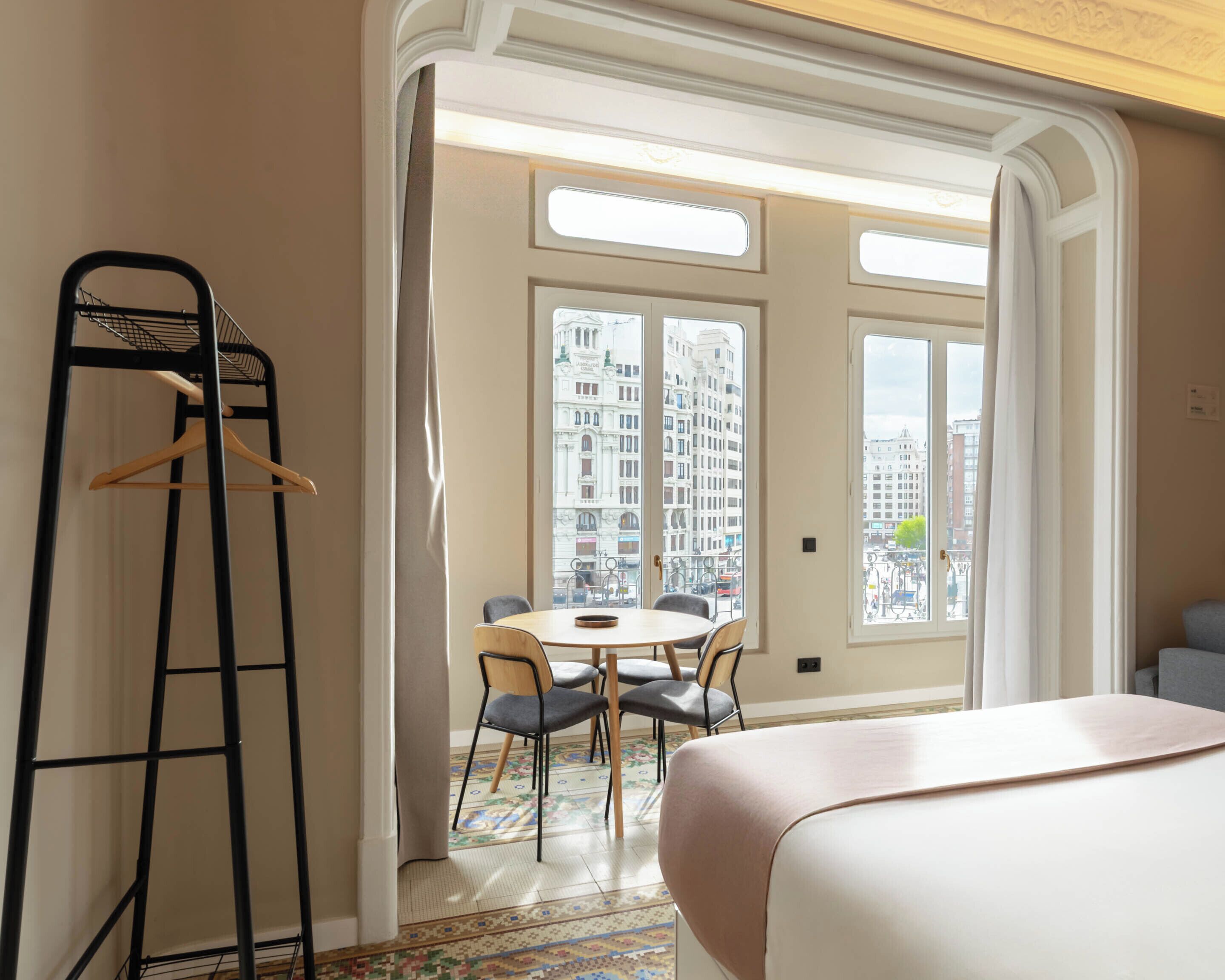 Camera d'albergo con un letto, un tavolo da pranzo con sedie, una rastrelliera in metallo nero e ampie finestre che offrono vista sulla città.