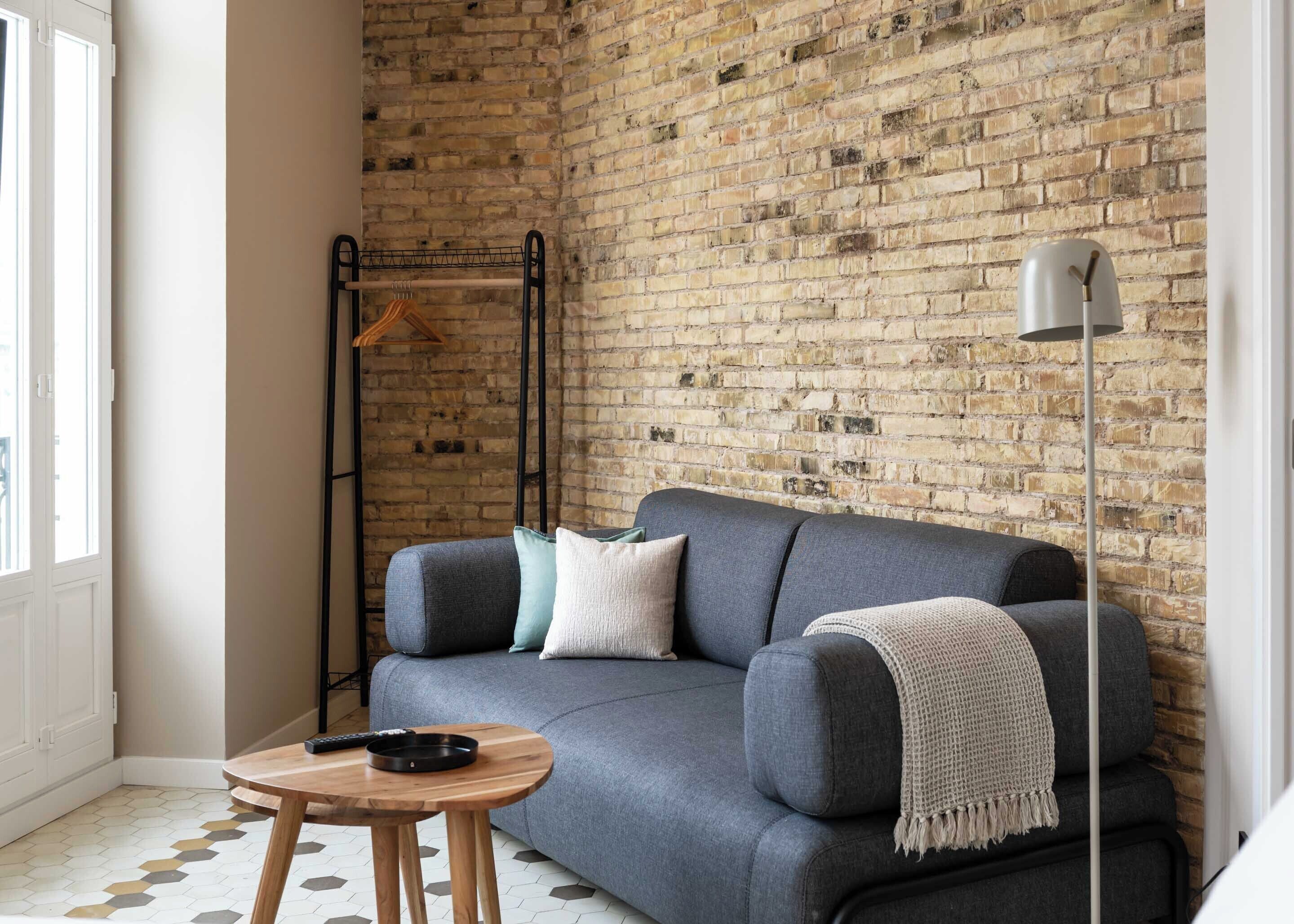 Una habitación con un sofá gris, una mesa de centro de madera, una pared de ladrillo, un perchero colgante y una lámpara de pie.