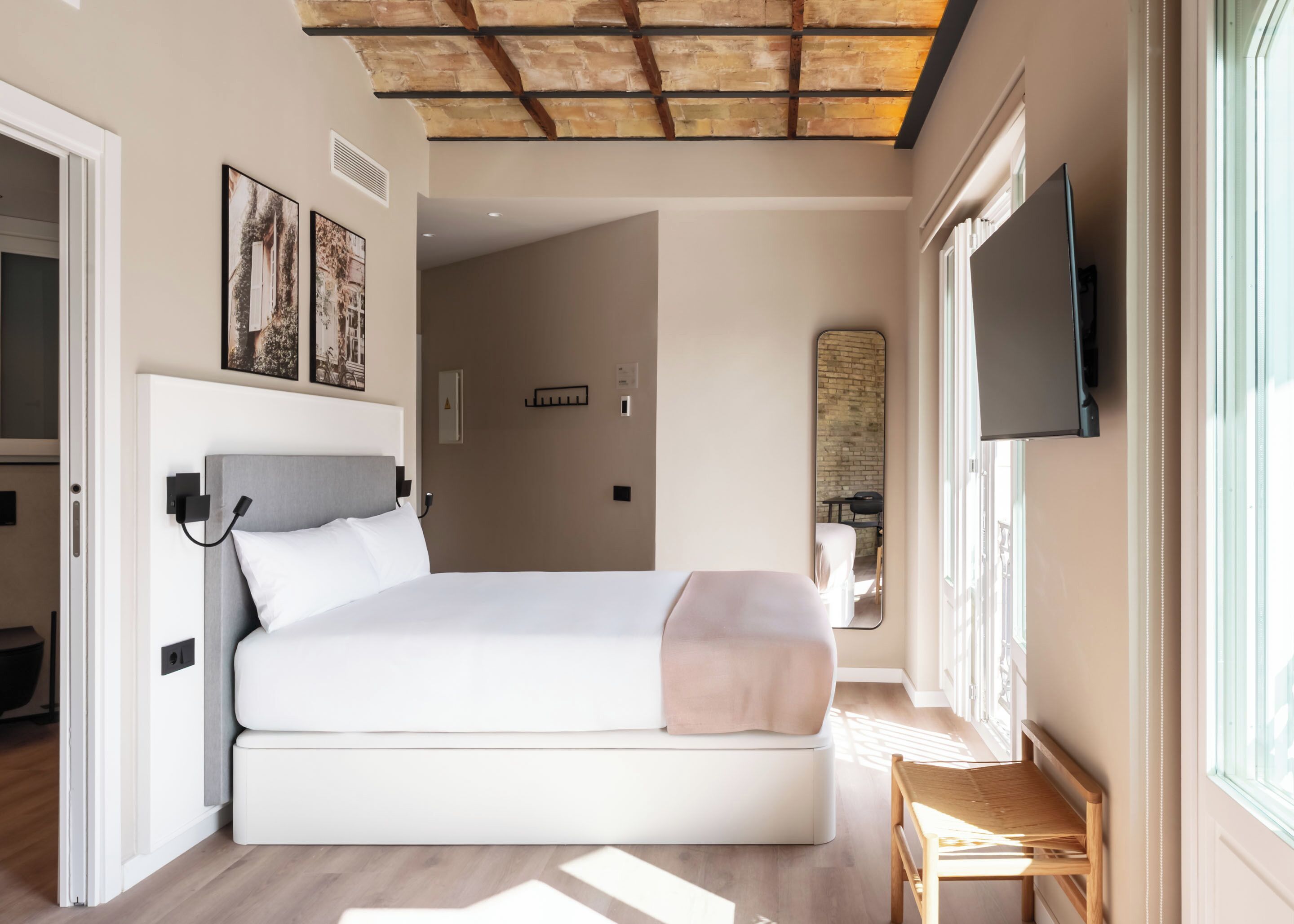 Ein modernes Hotelzimmer mit einem großen Bett, Deckenbalken aus Holz, einem an der Wand befestigten Fernseher und einem Fenster mit Tageslicht.