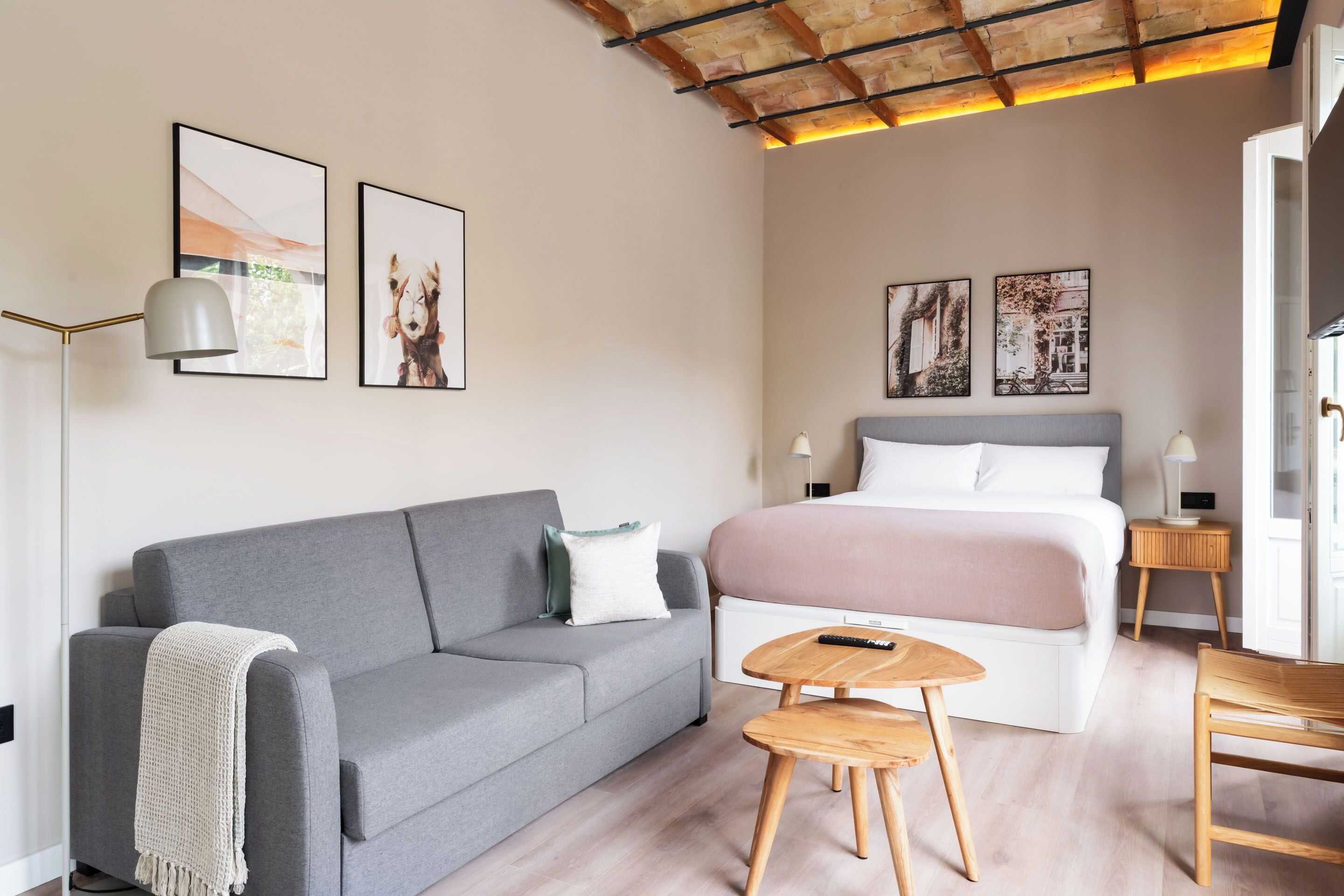 A modern bedroom with a grey sofa, a bed with a pink blanket, wooden side tables, and framed pictures on the wall.