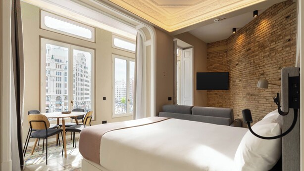 Une chambre d’hôtel dotée d’un grand lit, d’un canapé, d’une table à manger avec des chaises et offrant une vue sur la ville grâce à de grandes fenêtres.