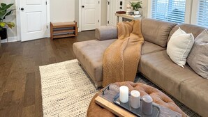 Un salón espacioso con un sofá de cuero marrón, una mesa de centro de madera con velas, un tapete blanco y un ventilador de techo.