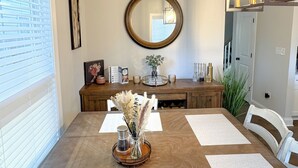 Un área de comedor con una mesa de madera, individuales blancos, un jarrón decorativo en el centro y un espejo redondo sobre la mesa.