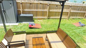 Un patio al aire libre con dos sillas de madera y una mesa, un área cubierta con techo de metal y una valla de madera al fondo.