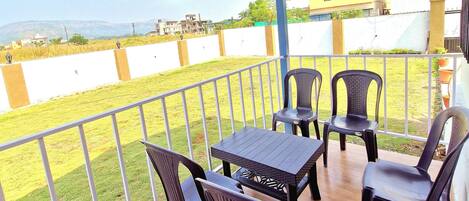 A covered outdoor seating area with a table and chairs, a hanging wind chime, and a view of a grassy field and buildings in the background.