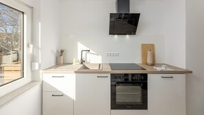 A modern kitchen with white cabinetry, a built-in oven, a stainless steel range hood, and a window providing natural light. - Troja Living - Apartment Helena am Park in the middle of Bruchsal (Bruchsal)