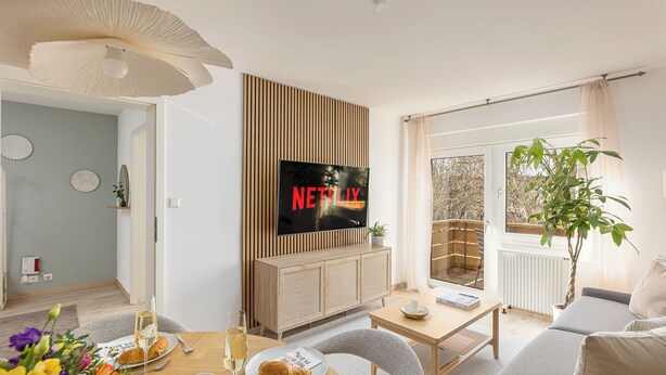 A modern living room with a flat-screen TV displaying 'NETFLIX', a dining area with a table set for two, and a large window with a view of the outdoors. - Troja Living - Apartment Helena am Park in the middle of Bruchsal (Bruchsal)