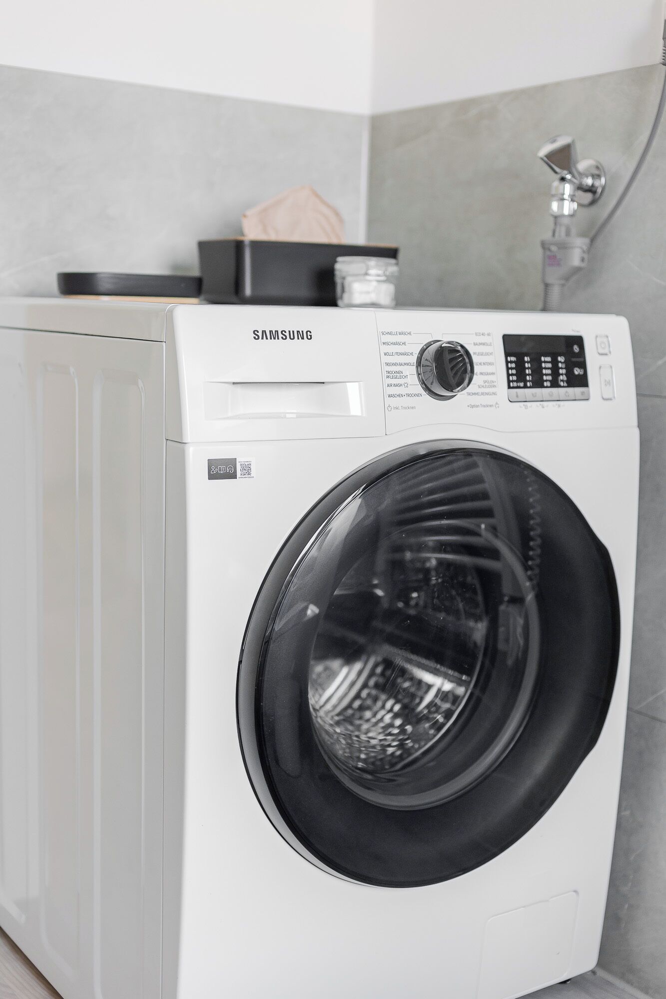 A Samsung front-loading washing machine in a bathroom with a tiled wall and a shower.