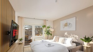 A modern bedroom with a large bed, a TV, a desk, and a view of the outdoors. - Troja Living - Apartment Helena am Park in the middle of Bruchsal (Bruchsal)