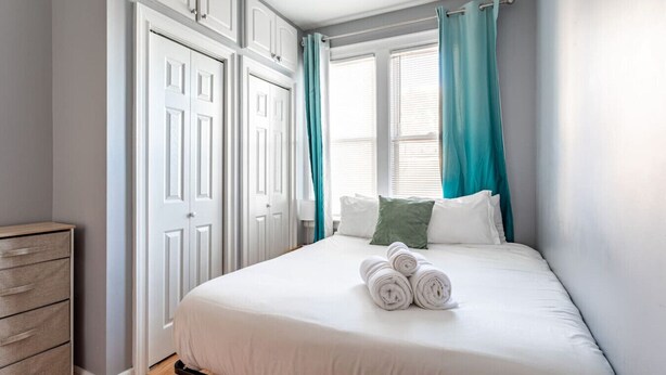 Une chambre à coucher avec un lit, des rideaux blancs, une commode en bois et une fenêtre avec des stores.