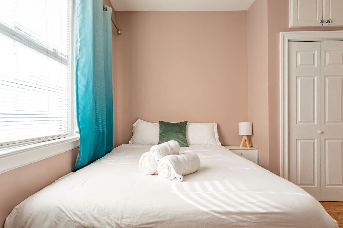 Een net opgemaakt bed met witte lakens en opgerolde handdoeken, een groen kussen en een bedlamp op een nachtkastje.