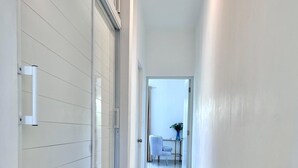 A modern hallway with a glass door, a white tiled floor, and a view into a room with a white chair and a vase.
