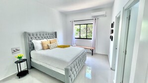 A modern bedroom with a tufted headboard, a large window with curtains, a bedside table with a vase of flowers, and a bench.