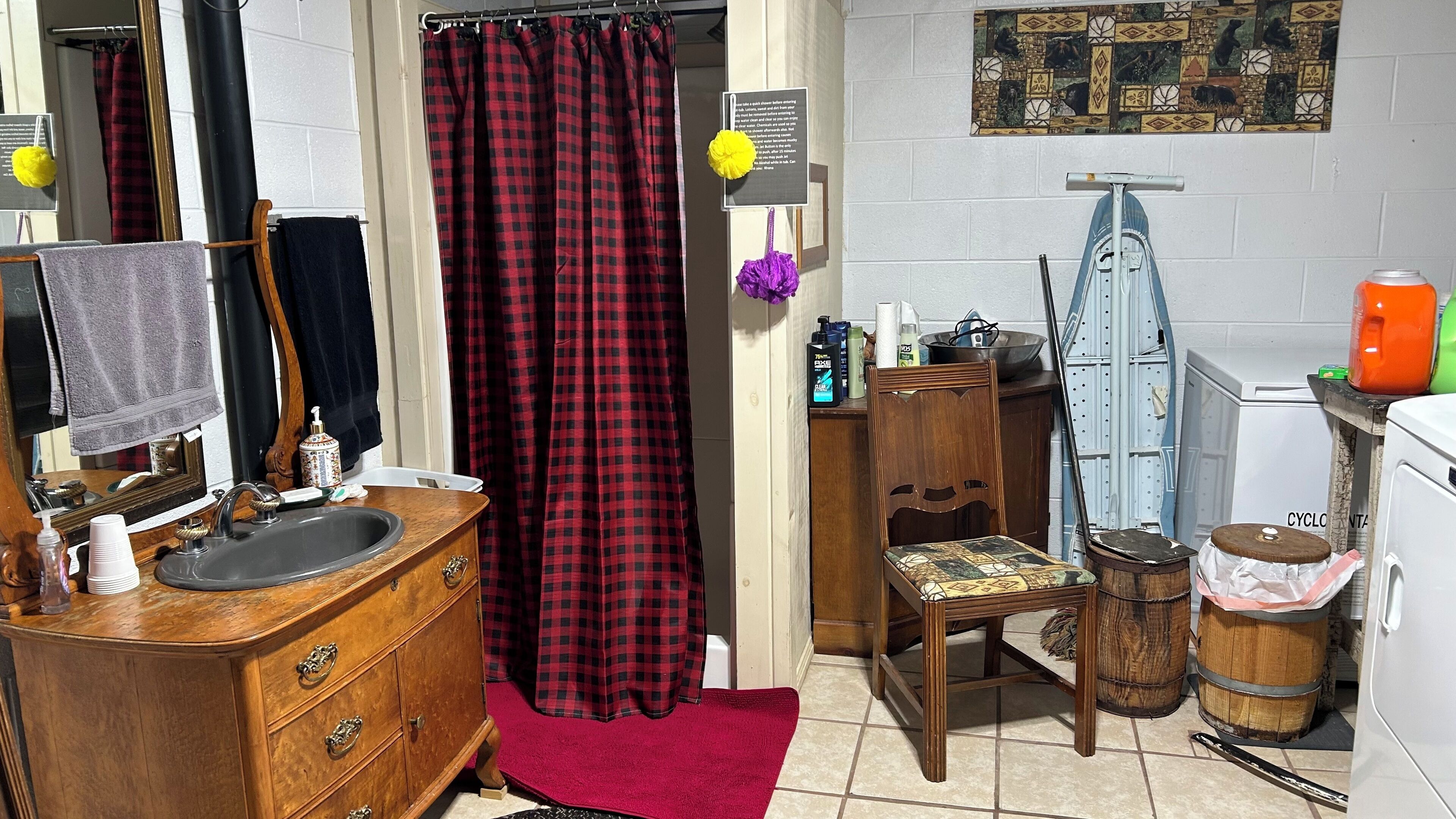 Une salle de bain avec un meuble-lavabo en bois, un miroir, un rideau de douche à carreaux rouges et noirs, une chaise en bois, une petite table avec des articles de toilette et un lave-linge blanc.