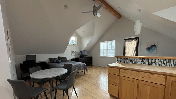 Een ruime loft slaapkamer met een schuine kap, een eethoek met een ronde tafel en stoelen, en een keuken met houten kastjes en een betegelde spatwand.