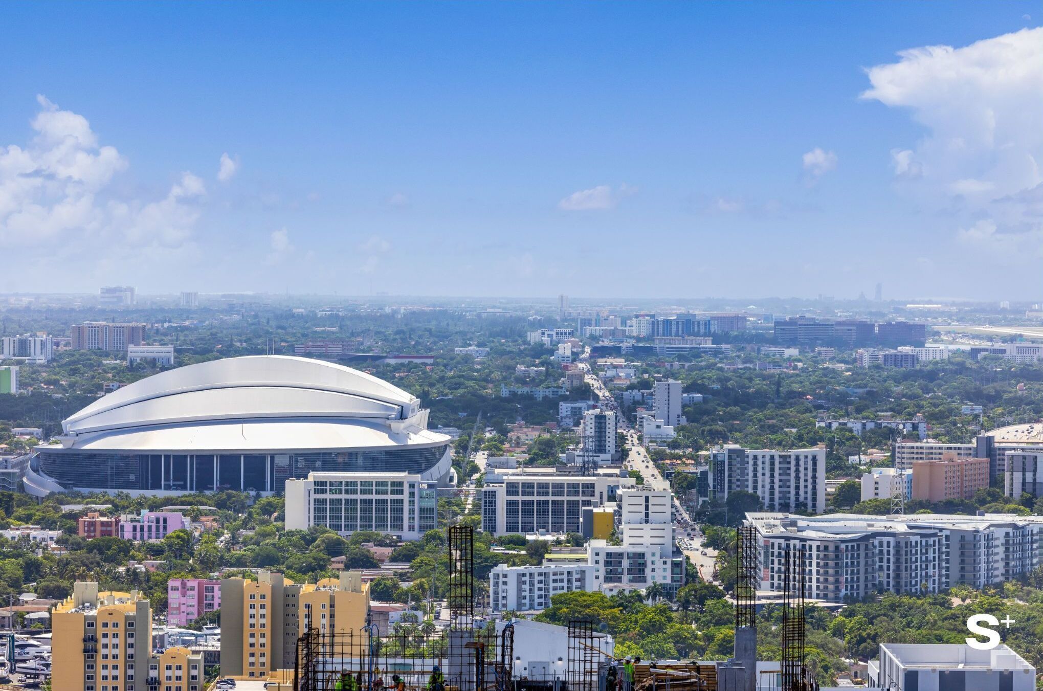 A modern stadium with a large, curved roof, surrounded by urban buildings and greenery.