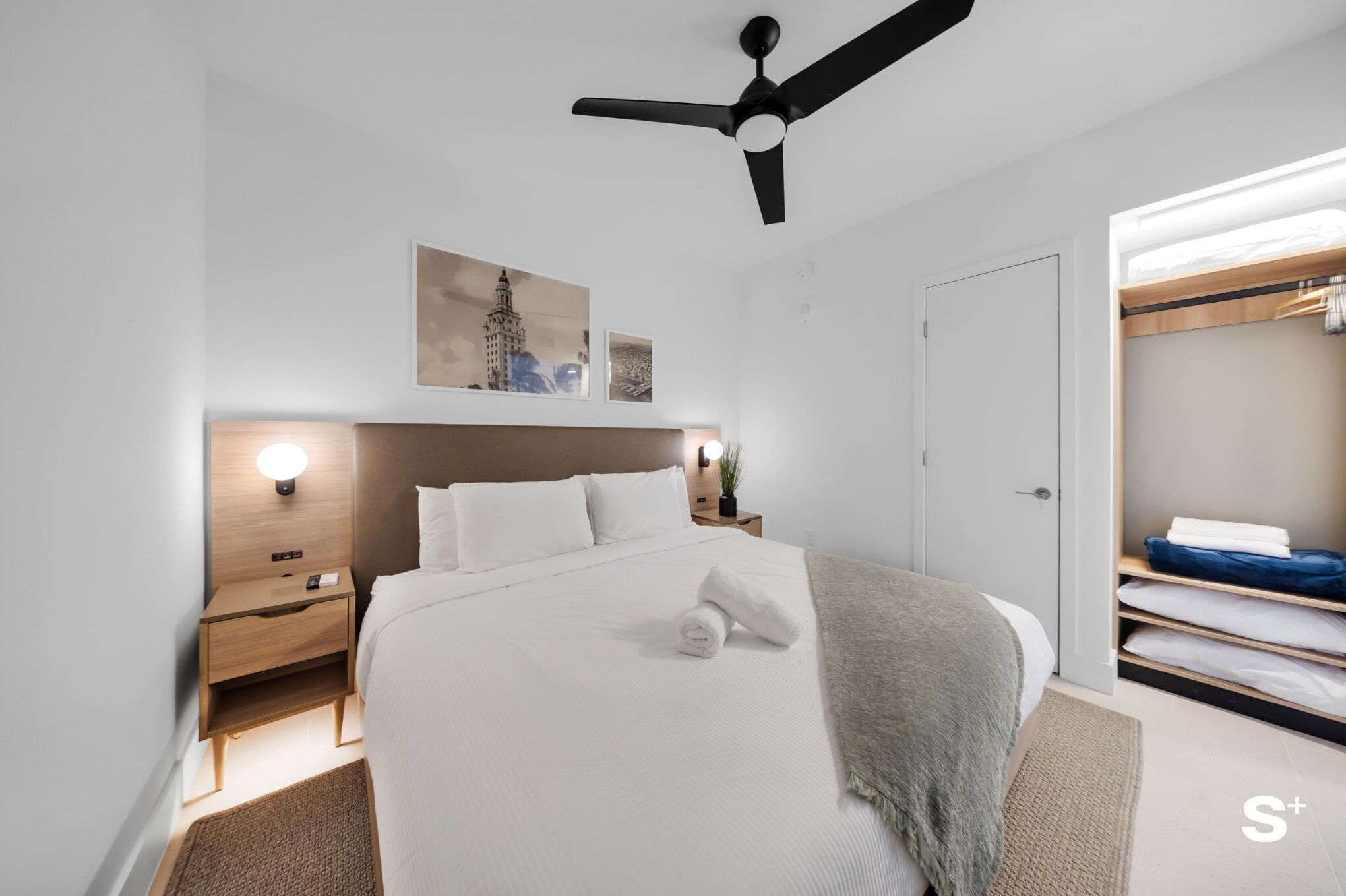 A modern hotel room with a neatly made bed, a ceiling fan, a bedside table with a lamp, and a built-in closet with hanging space.