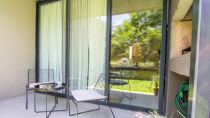 A balcony with two chairs and a table, a large window with curtains, and a view of a garden and trees. - Premium Depto con jardin Palermo C1A (Buenos Aires)