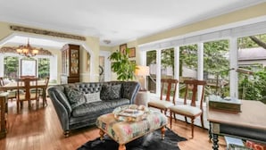A spacious living room with a dark leather sofa, a floral ottoman, and wooden chairs. A dining table is visible in the background.