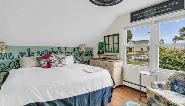 Et soveværelse med en stor seng, en kommode, en stol og et vindue med udsigt til et hus og grønne områder.