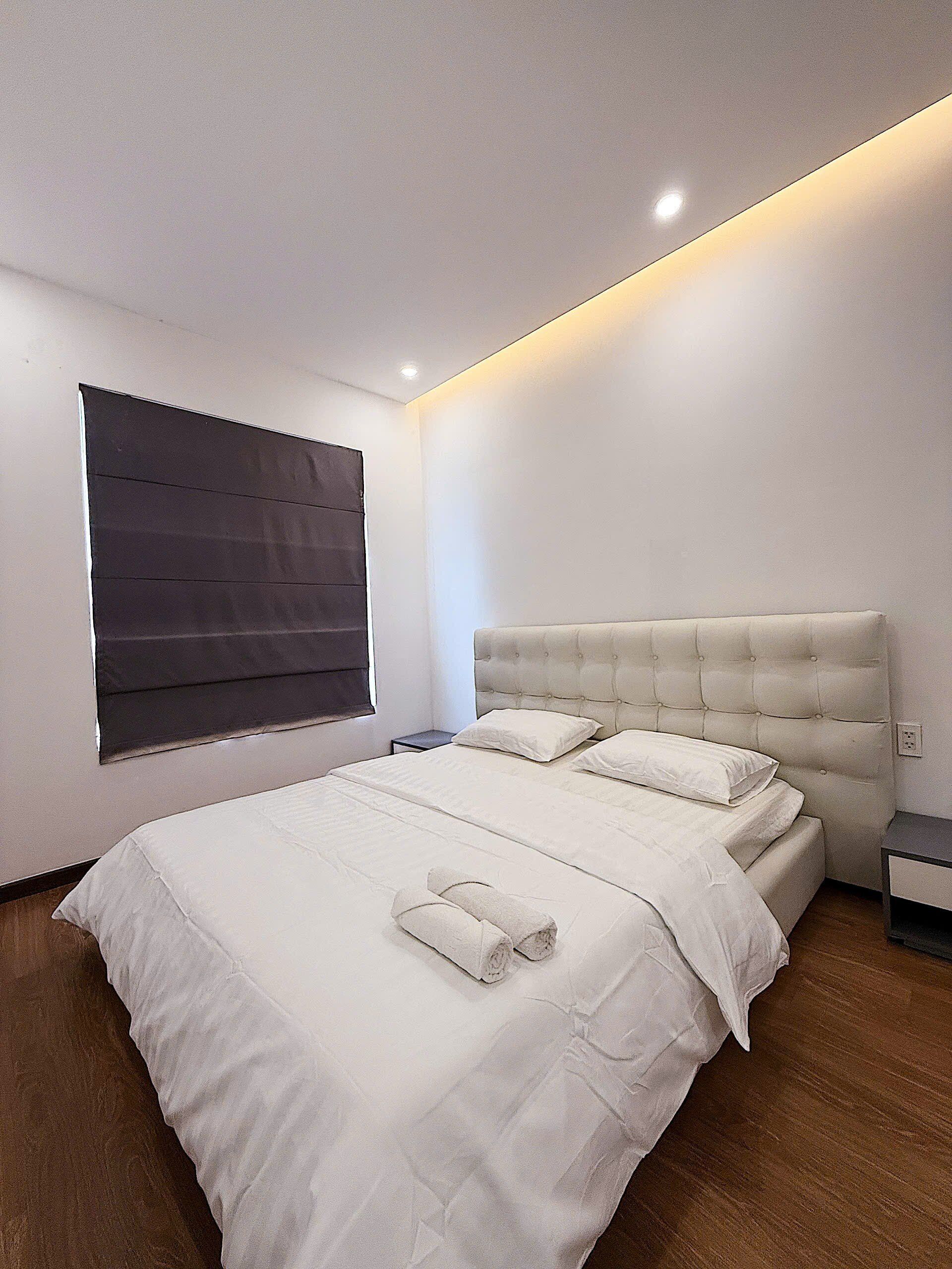 A modern bedroom with a large bed, a bedside table, and a window with blinds.