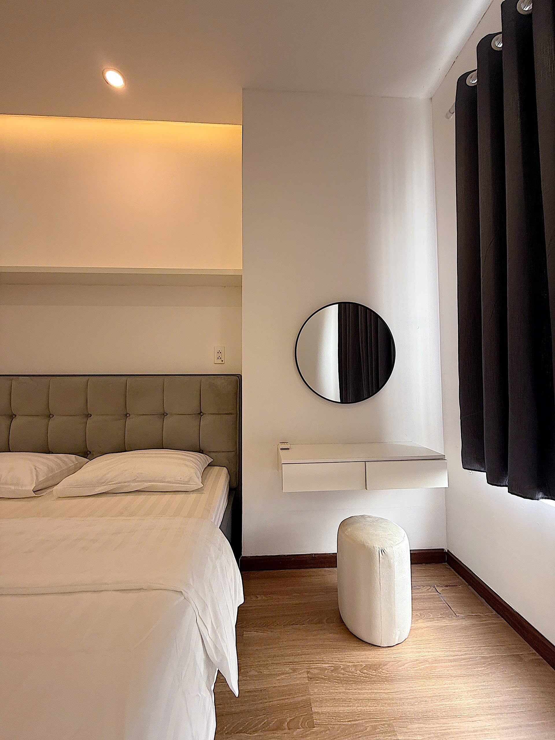 A bedroom with a bed, a round mirror, a white bedside table, and a window with curtains.