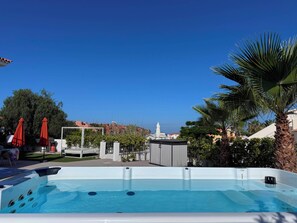 Une piscine avec un jacuzzi, entourée de palmiers et de parasols rouges, d’où l’on aperçoit des bâtiments en arrière-plan.