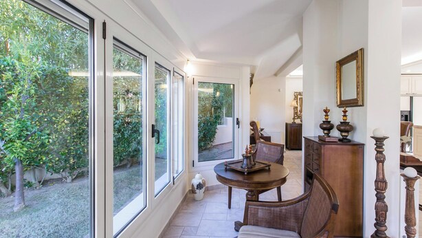 Una habitación amplia con grandes puertas correderas de vidrio que dan a un jardín, una mesa redonda de madera con sillas y un armario de madera con adornos.