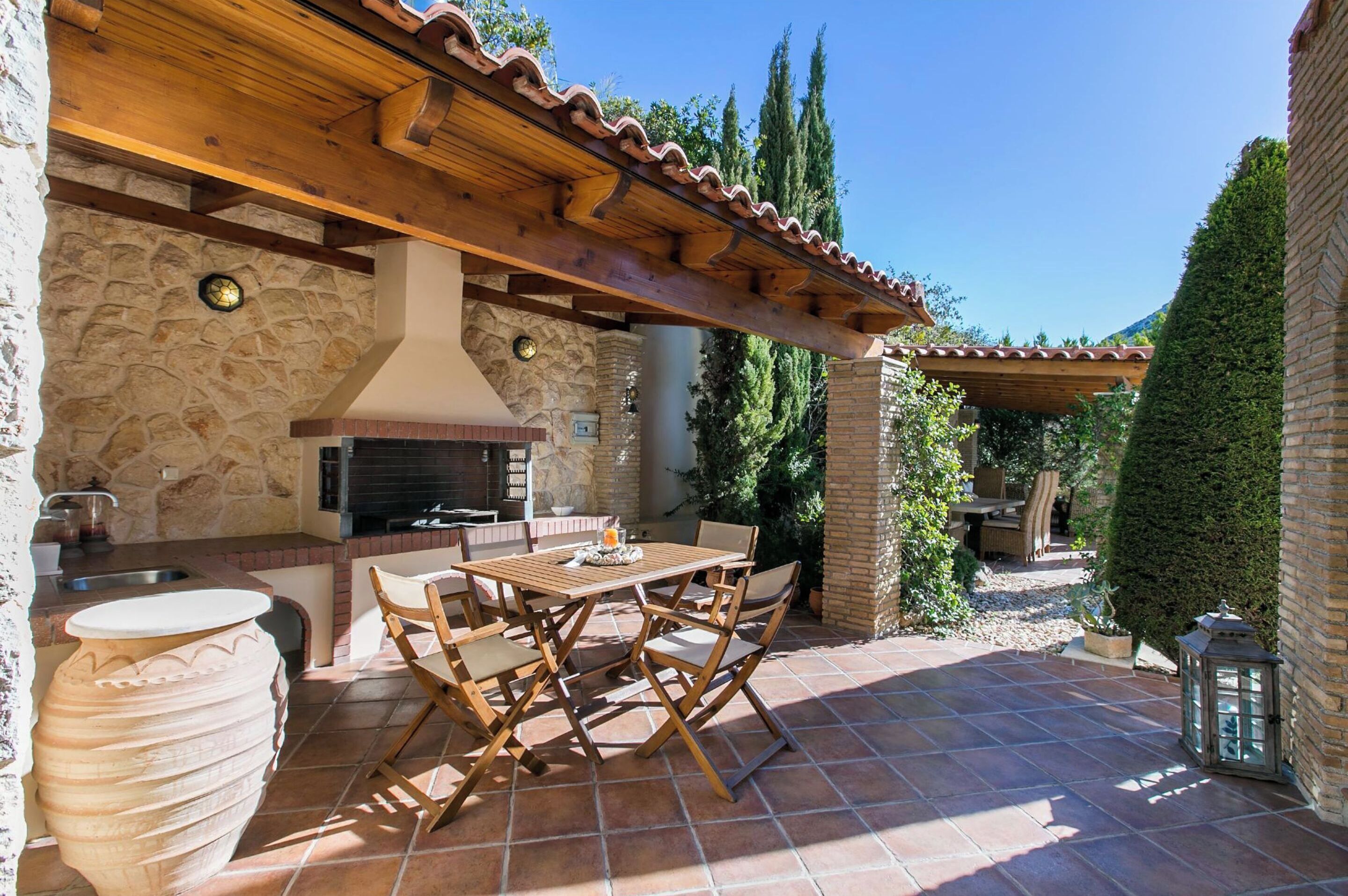 Un área de cocina al aire libre con pared de piedra, pérgola de madera y una mesa con sillas.