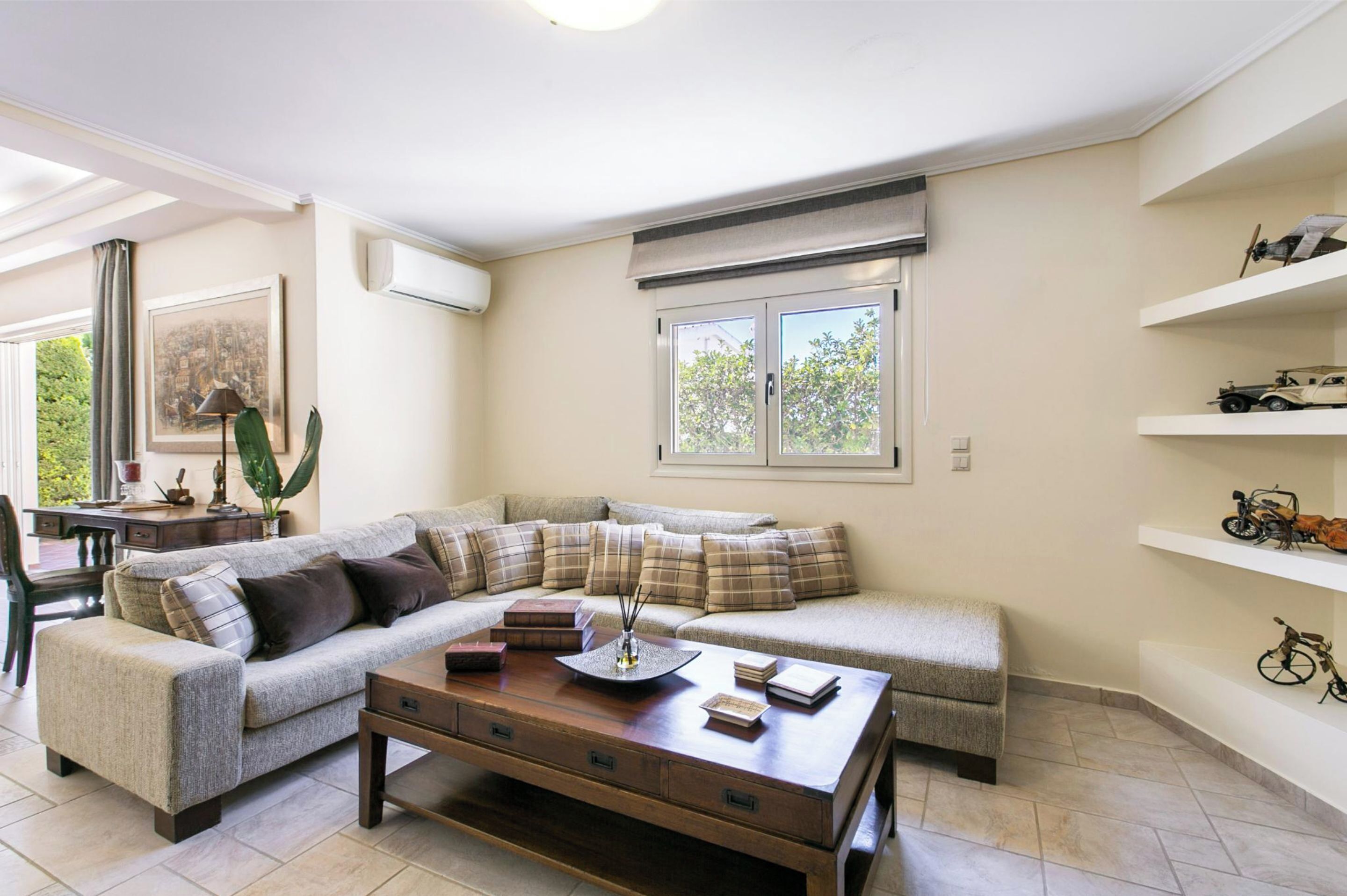 A living room with a sectional sofa, a coffee table, and a desk with a lamp. There are shelves with decorative items and a window with a view of trees.