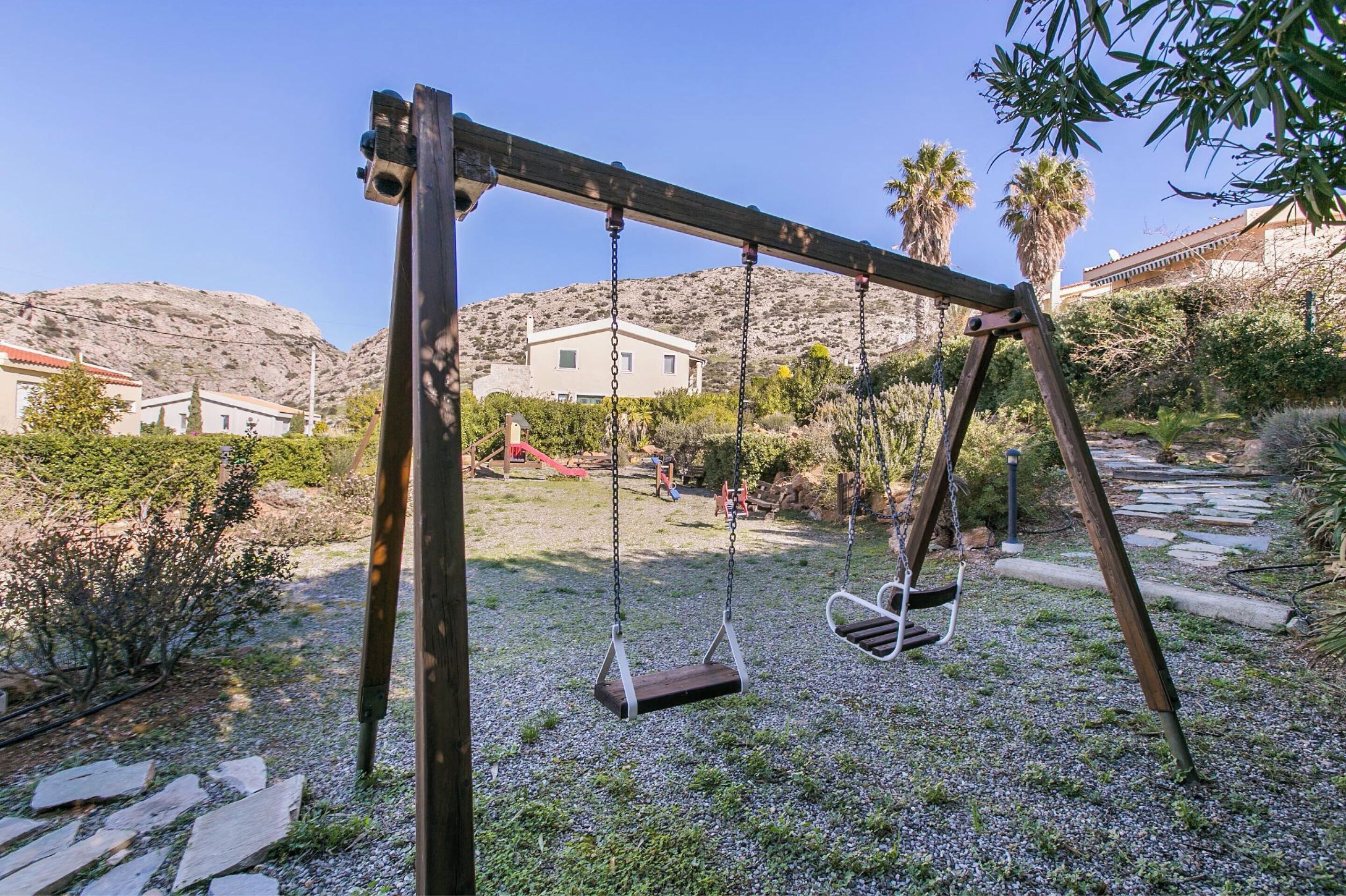 A wooden swing set in a garden with a stone pathway, surrounded by greenery and a mountain in the background.