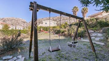 A wooden swing set in a garden with a stone pathway, surrounded by greenery and a mountain in the background.