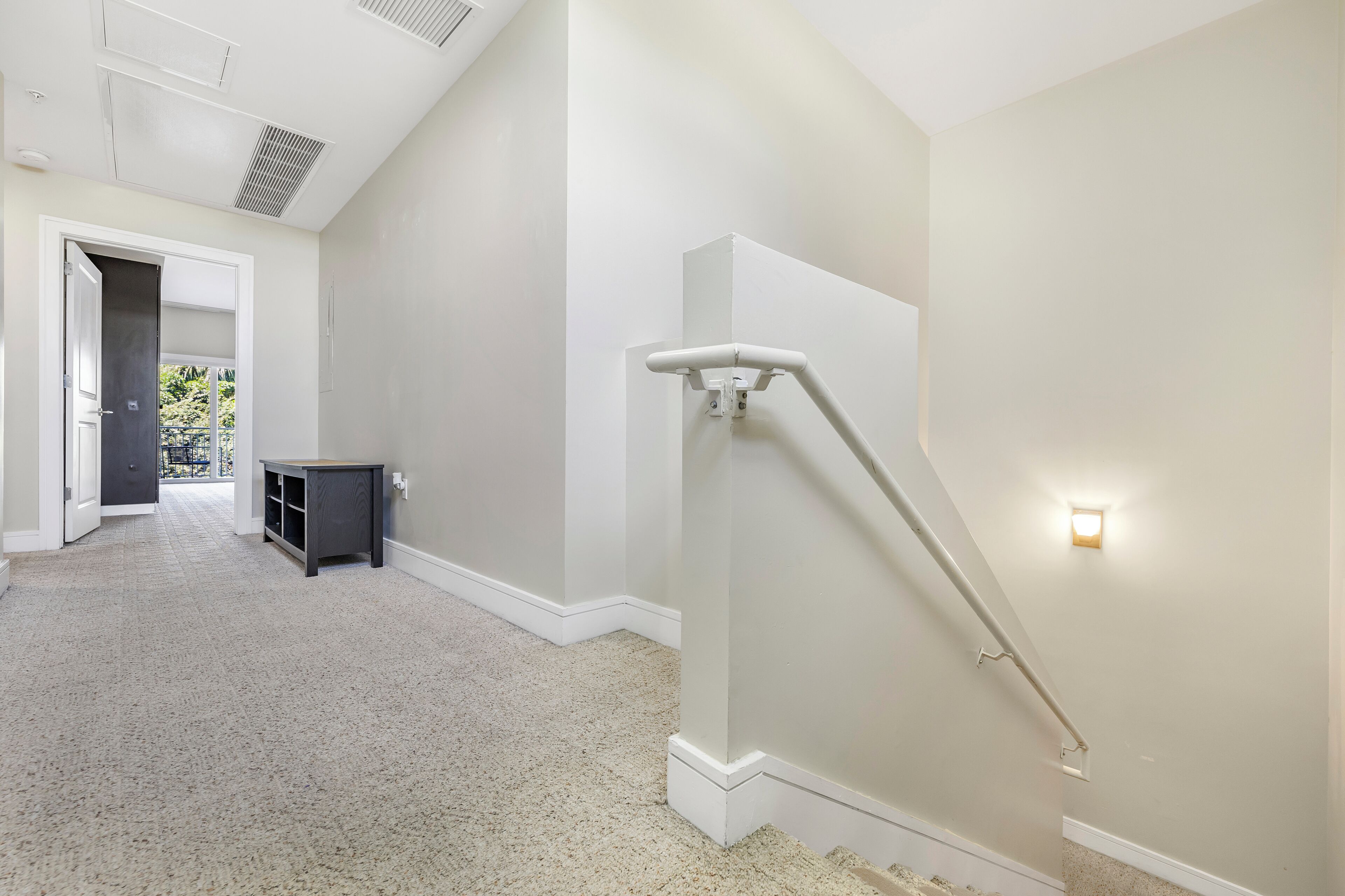 A hallway with a carpeted floor, a dark-colored bench, and a white wall with a light fixture.