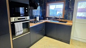 A modern kitchen with dark cabinetry, a herringbone-patterned floor, a built-in oven, and a window overlooking a residential area. - Quirky 2-Bed with Free Parking (Paisley)