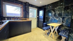 A modern kitchen with a central island, a dining area with a wooden table and blue upholstered chairs, and a large window providing natural light. - Quirky 2-Bed with Free Parking (Paisley)