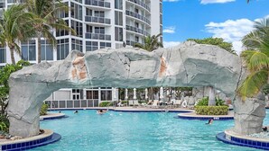 Una piscina con un’imponente arcata rocciosa, palme e un edificio sullo sfondo.