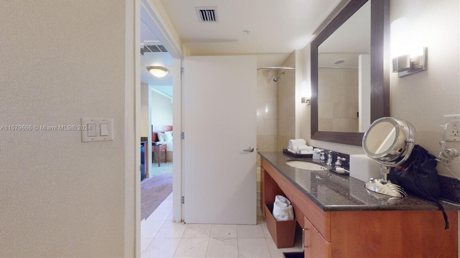 A bathroom with a wooden vanity, a large mirror, and a shower area visible through an open door.