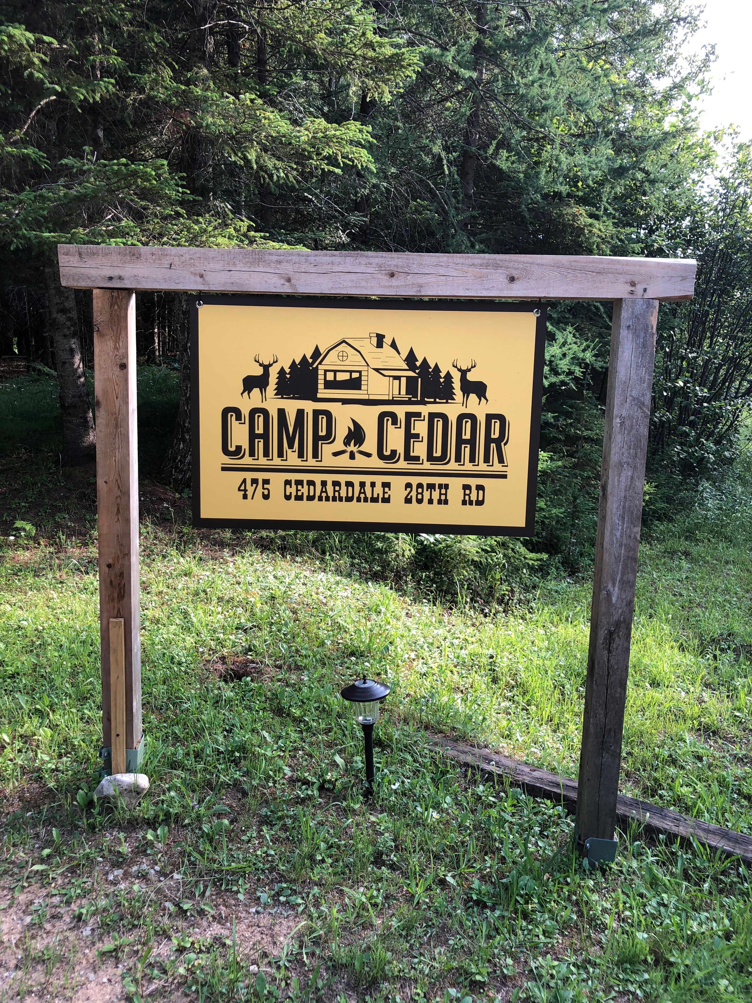 Een houten bord met de tekst 'Camp Cedar 475 Cedardale 28th Rd' en een afbeelding van een blokhut met herten.
