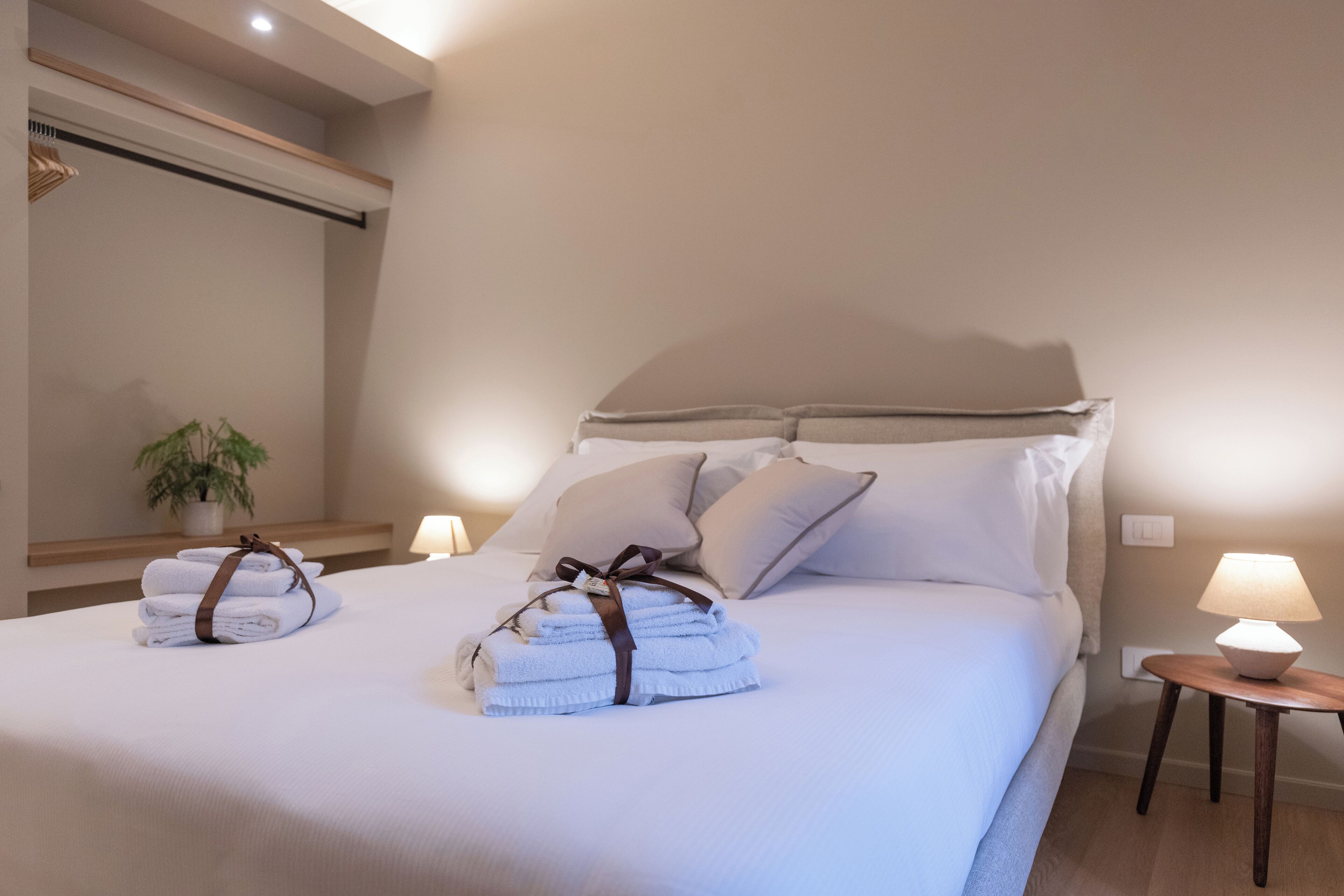 A neatly made bed with towels and pillows, a bedside table with a lamp, and a potted plant on a nightstand.