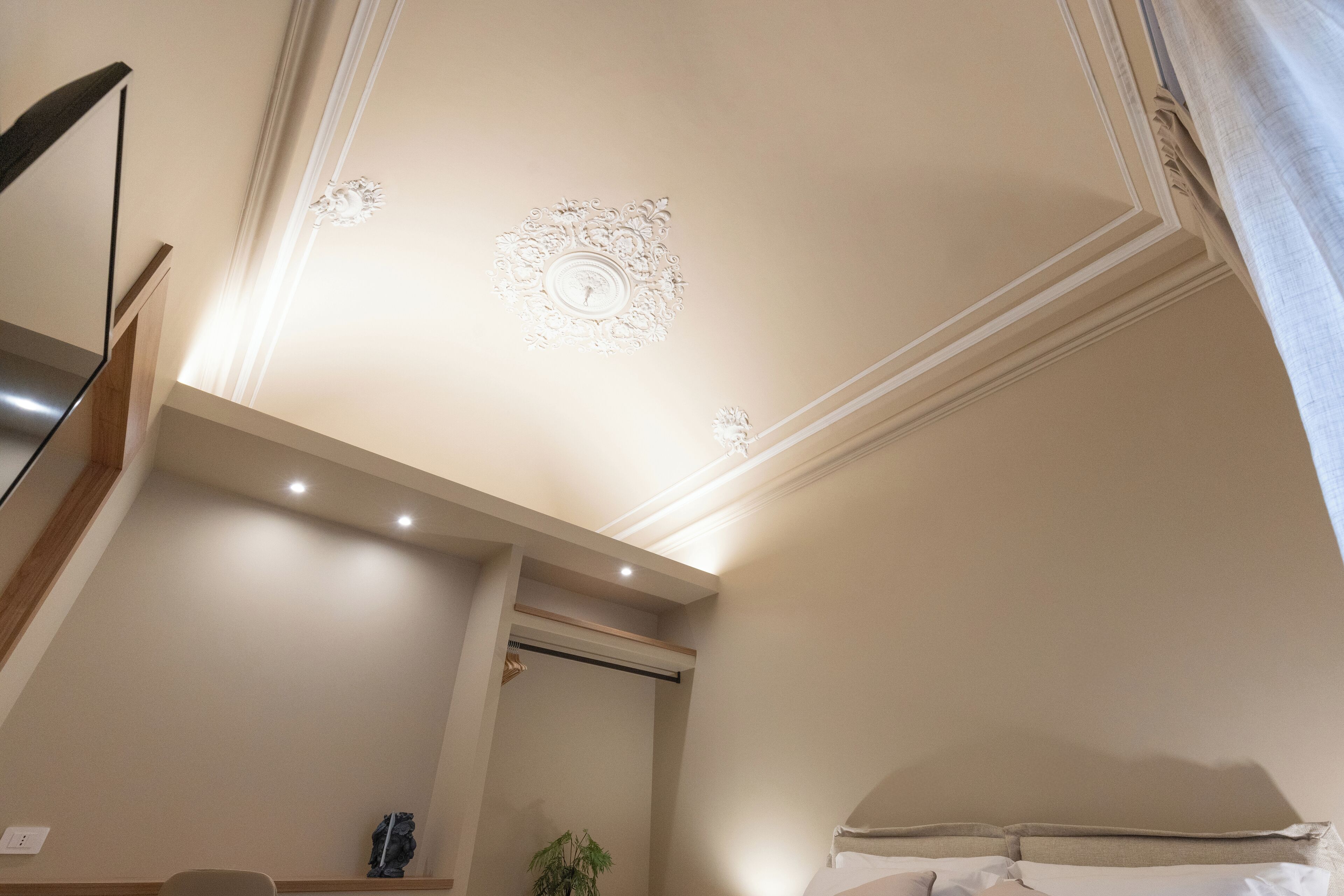 Ein Hotelzimmer mit Bett, Nachttisch, Spiegel, Pflanze und einer Decke mit dekorativen Stuckverzierungen.