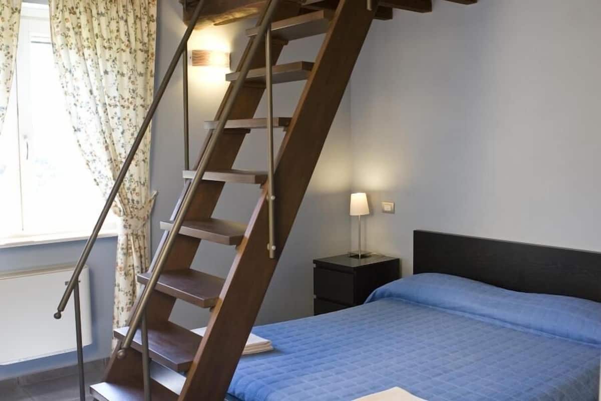 A bedroom with a wooden staircase, a bed with a blue quilt, a nightstand with a lamp, and a window with floral curtains.