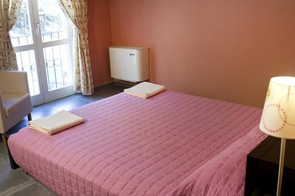 A room with a pink bedspread, a chair, a lamp, and a window with curtains.