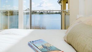 A bedroom with a large window overlooking a body of water, a bed with a book, and a balcony with a railing. - Squirrels Leap: Lake View with Spa Access (Somerford Keynes)