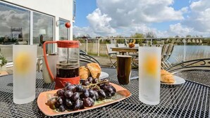 A table with a coffee maker, croissants, grapes, and glasses of water, set outdoors with a view of a building and a body of water. - Squirrels Leap: Lake View with Spa Access (Somerford Keynes)