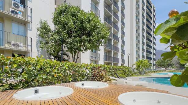 A wooden deck with hot tubs, a pool, and a building with balconies in the background. - Modern Apartment with a Spectacular Ocean View (Santa Marta)