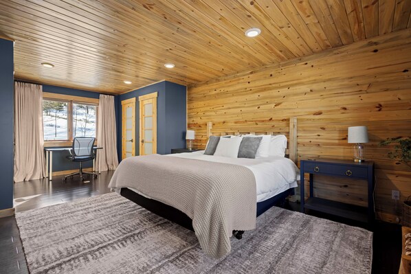 Un dormitorio con techo de madera, cama con ropa de cama blanca, escritorio con silla y ventana con cortinas.
