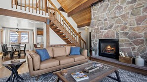 Un salon chaleureux avec une cheminée en pierre, un escalier en bois, un canapé beige agrémenté de coussins bleus et une table basse avec des magazines.