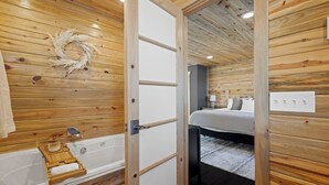 L'intérieur d'un chalet en bois, avec une baignoire en bois, un lit recouvert d'une couverture grise et un mur blanc orné d'une guirlande décorative.