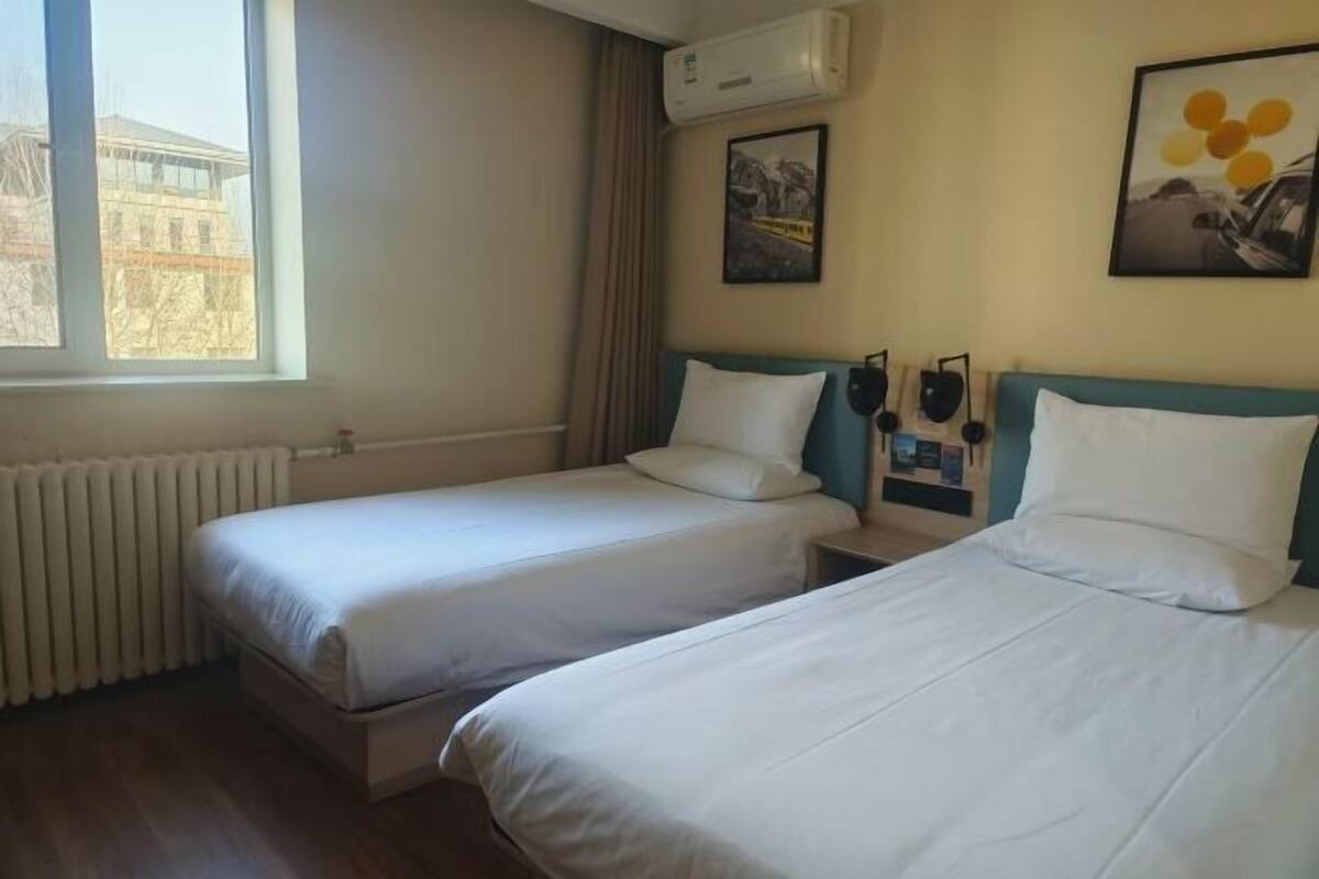 A hotel room with two single beds, a window with a view of buildings, an air conditioner, and a wall-mounted clock.