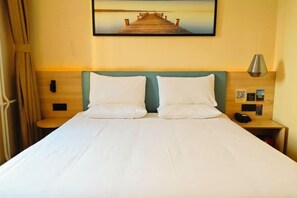Une chambre d’hôtel avec un lit, deux oreillers, une table de chevet avec une lampe, un tableau représentant une jetée et une tête de lit en bois.